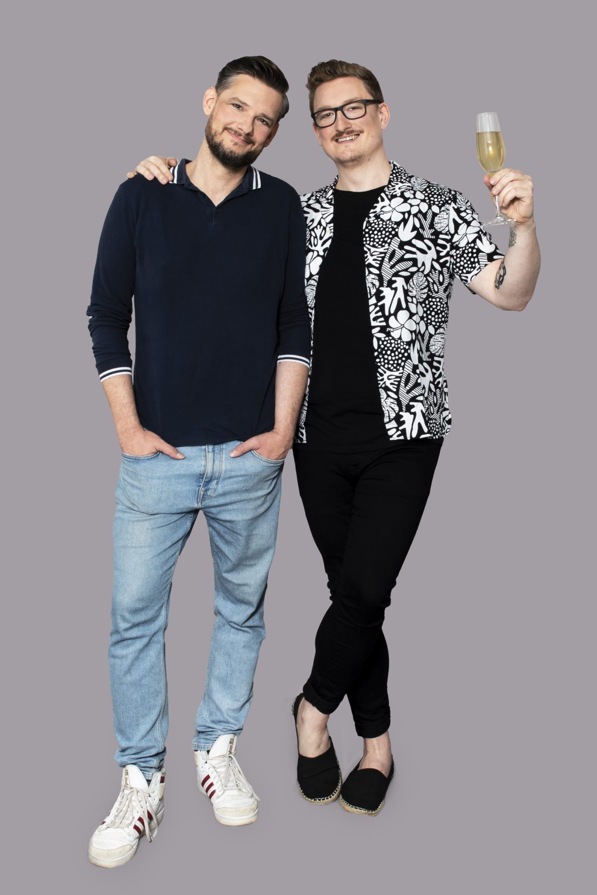 Bild zeigt zwei Personen. Eine Person hält ein Sektglas in der Hand und umarmt die andere Person mit dem rechten Arm