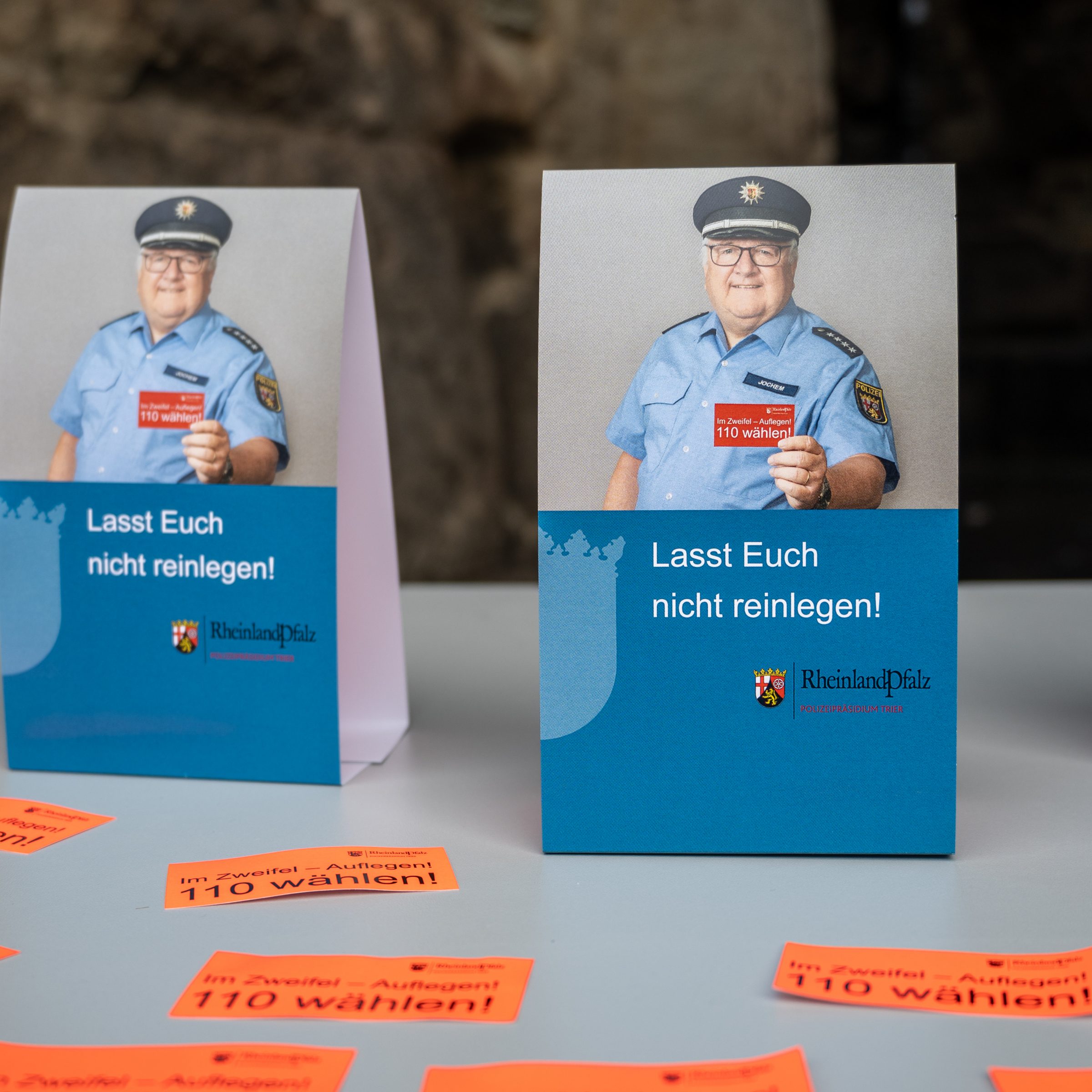 Zwei Aufsteller unseres Plakats, das einen Polizisten abbildet, der eine Karte in die Kamera hält, auf der "Im Zweifelsfall 110 wählen" steht. Als Titel des Plakates unter dem Foto steht "Lasst Euch nicht reinlegen!". Davor liegen auf einem Tisch mehrere rote Zettel auf denen "Im Zweifel- Auflegen! 110 wählen!" steht.