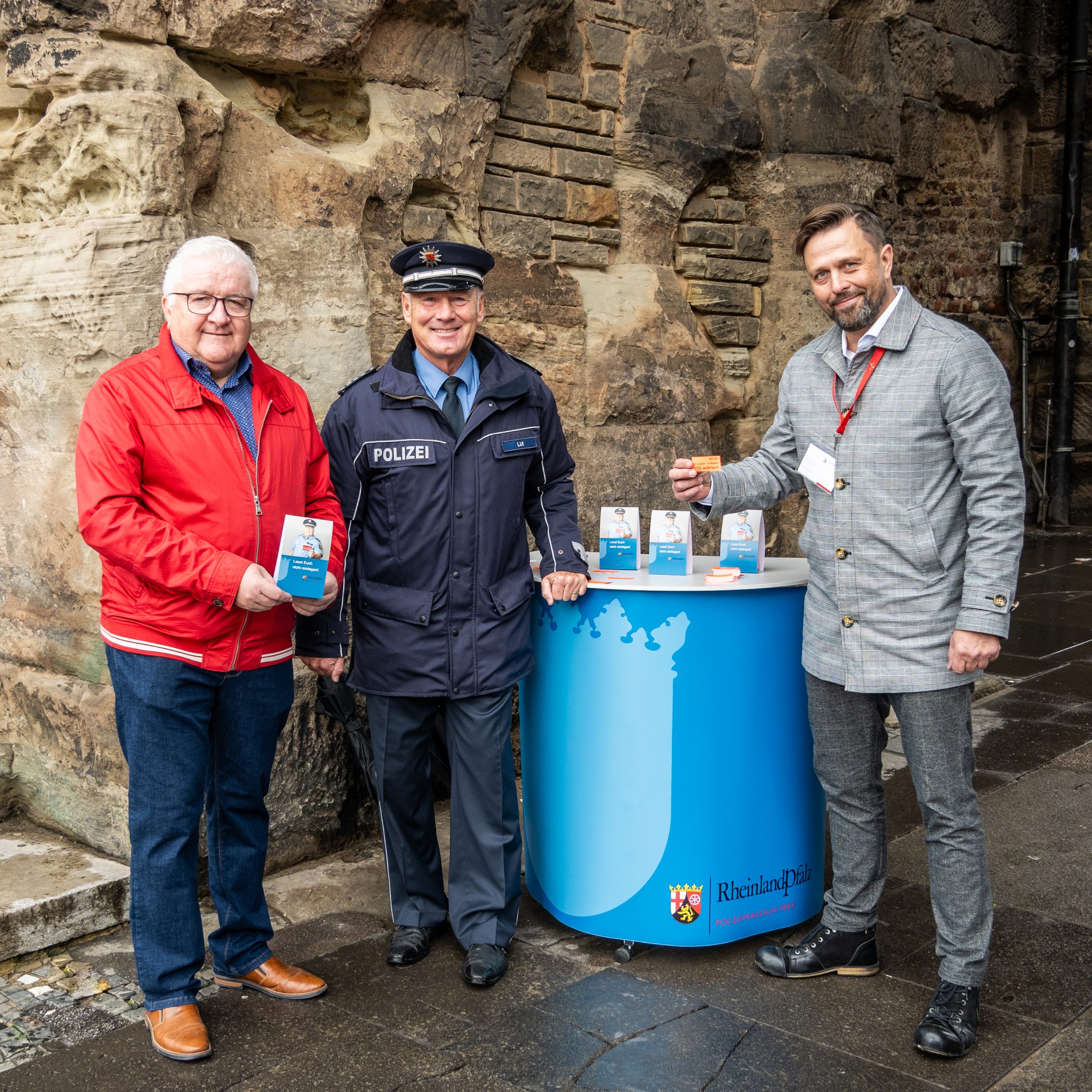 Drei Polizisten stehen nebeneinander um einen kleinen Stand herum, auf dem Aufsteller des Plakates stehen. Einer von ihnen hält eine rote Karte mit Schriftzug.