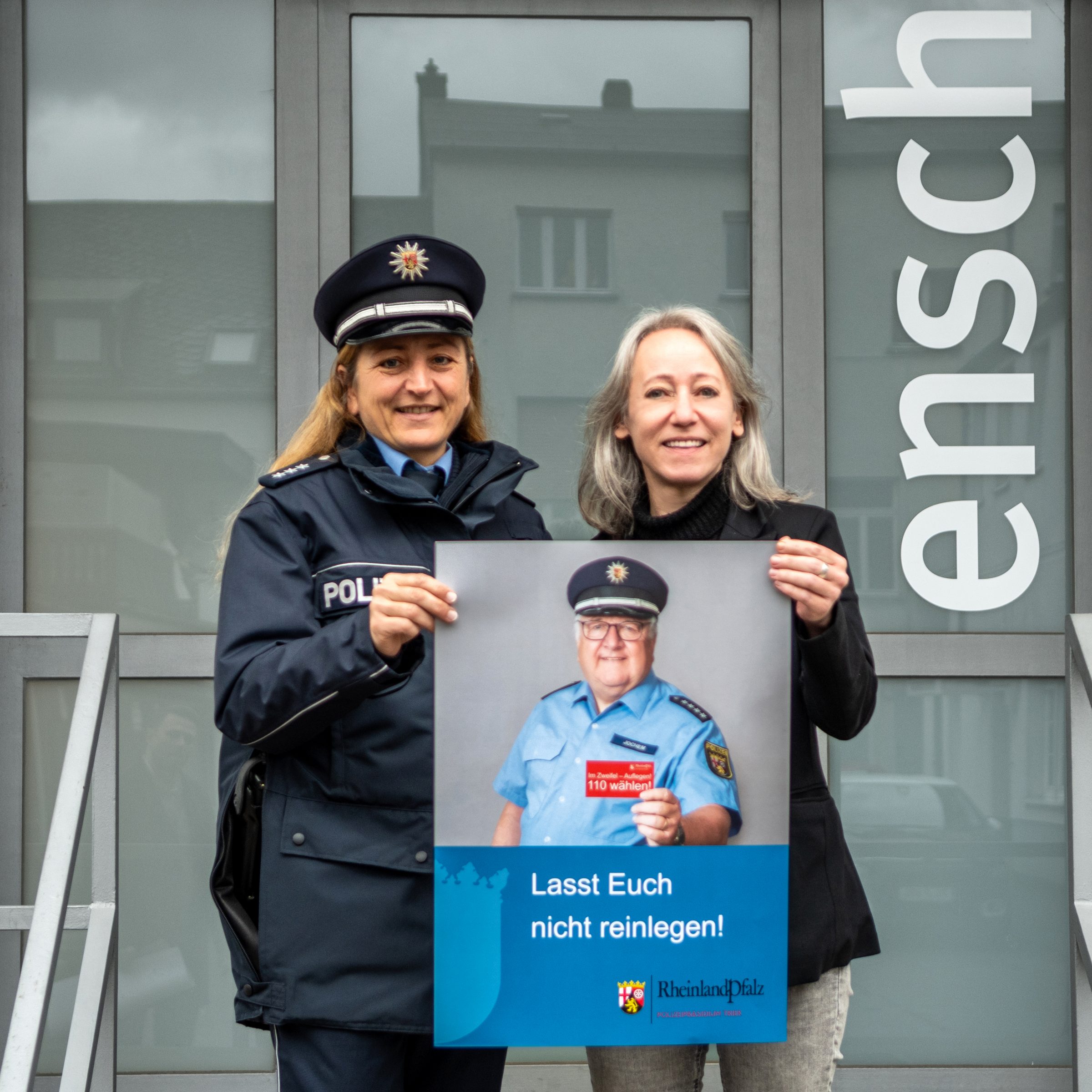 Eine Polizistin und ensch-media-Chefin halten zusammen ein Plakat, das einen Polizisten abbildet, der eine Karte in die Kamera hält, auf der "Im Zweifelsfall 110 wählen" steht. Als Titel des Plakates unter dem Foto steht "Lasst Euch nicht reinlegen!"