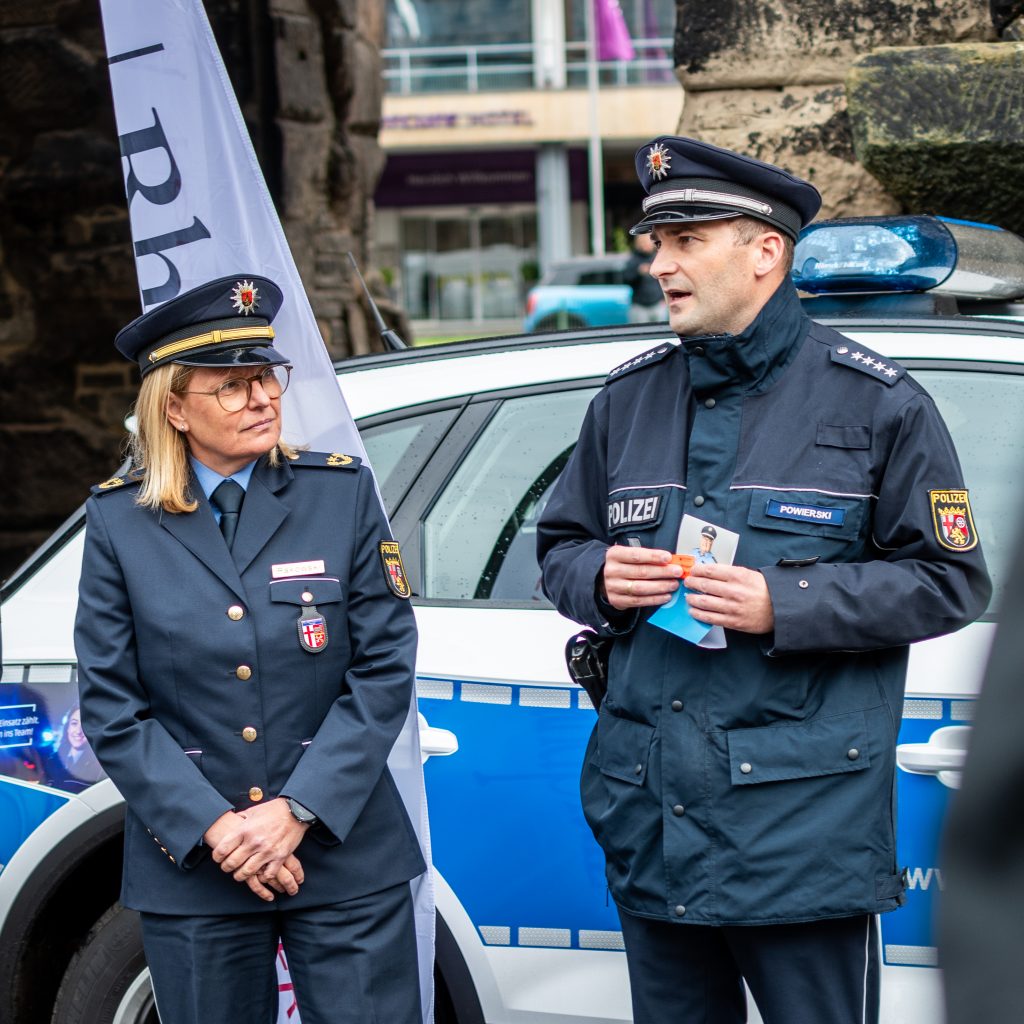 Ein Polizist und eine Polizistin stehen nebeneinander vor einem Polizeiauto, das an der Porta Nigra steht. Er redet.