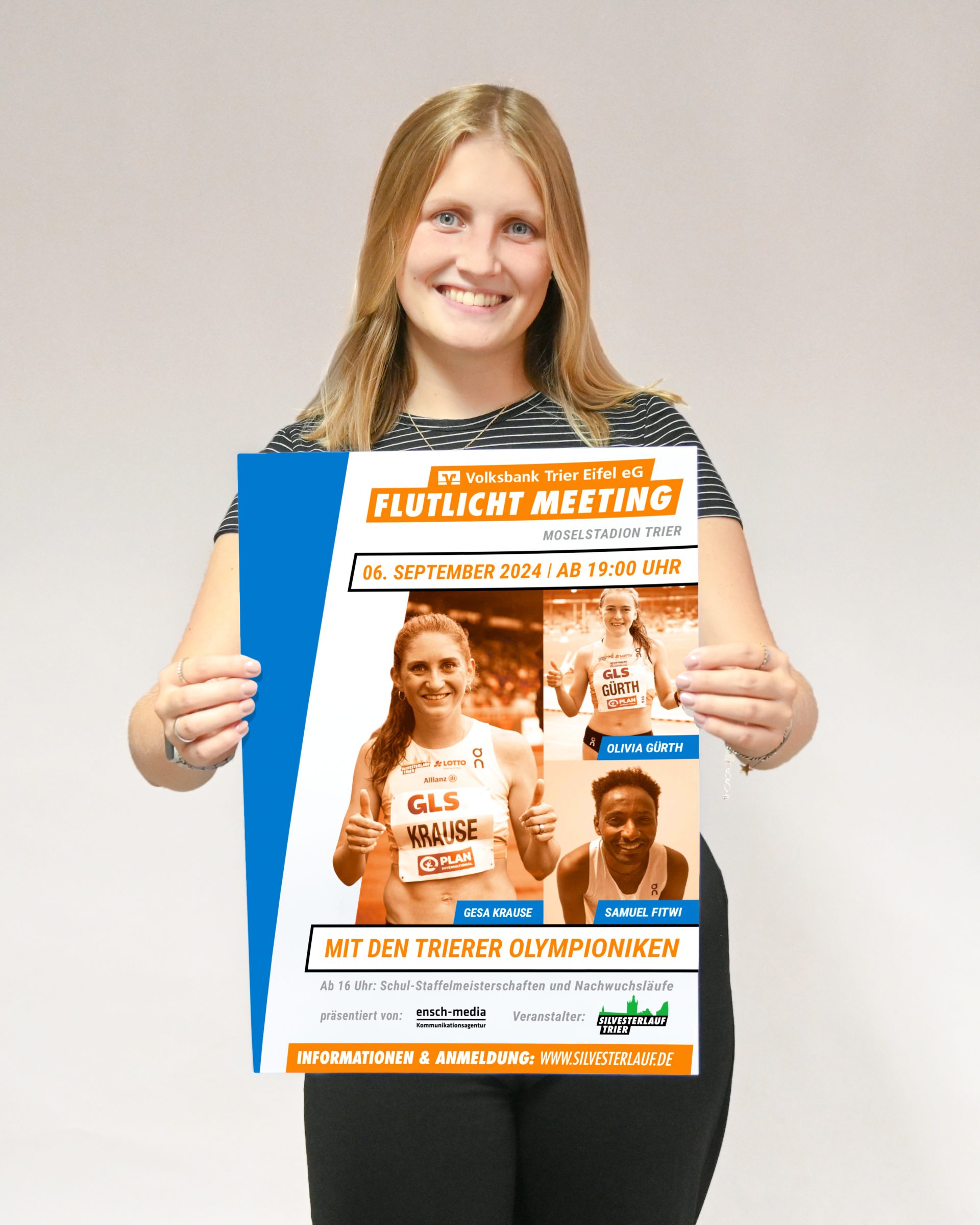 Frau lächelt und hält Plakat für das Volksbank-Trier Eifel-Flutlichtmeeting im Moselstadion Trier.