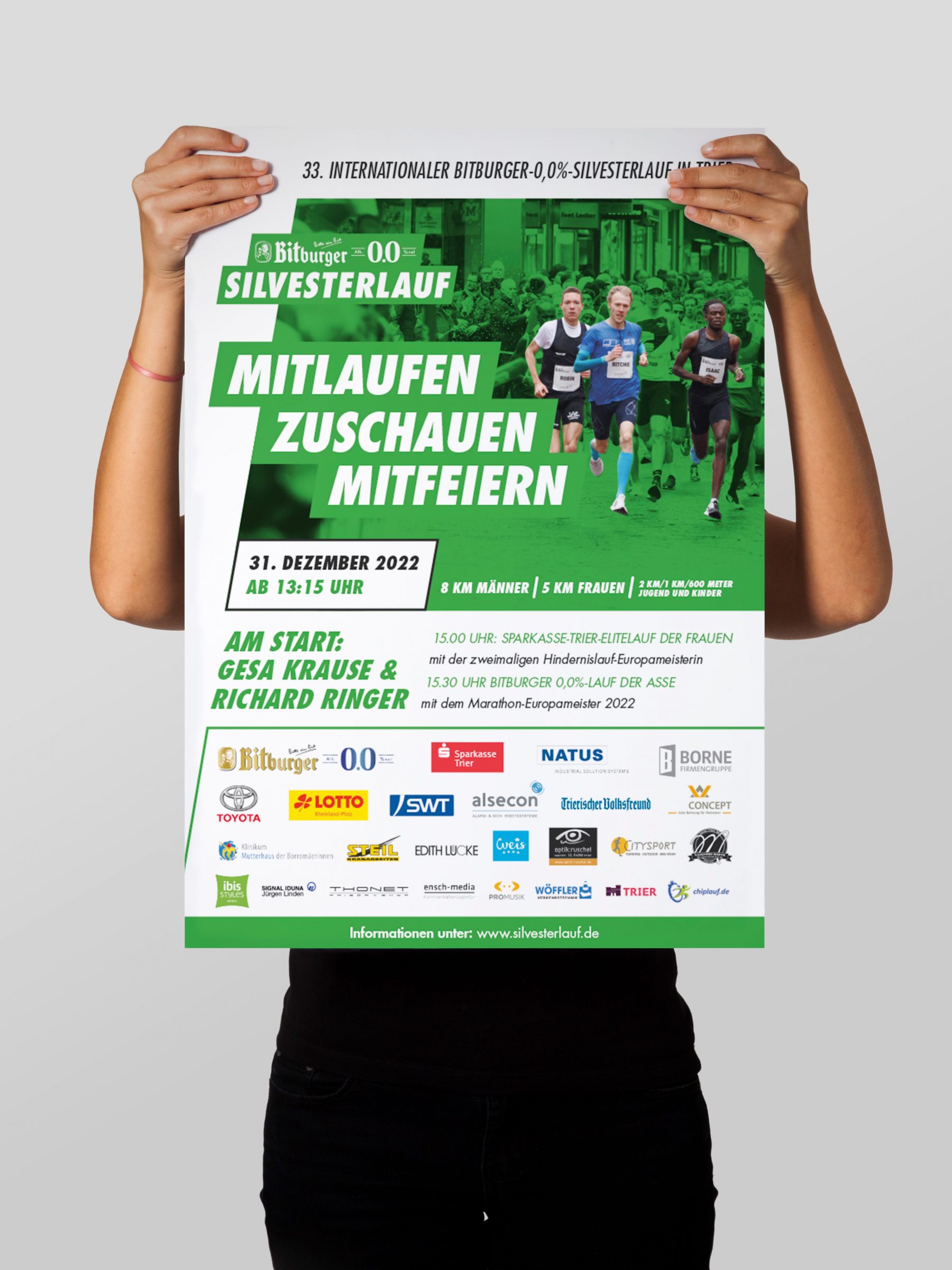 Eine Person hebt das Plakat des Bitburger 0,0%-Silvesterlaufs in Trier von 2022 hoch.