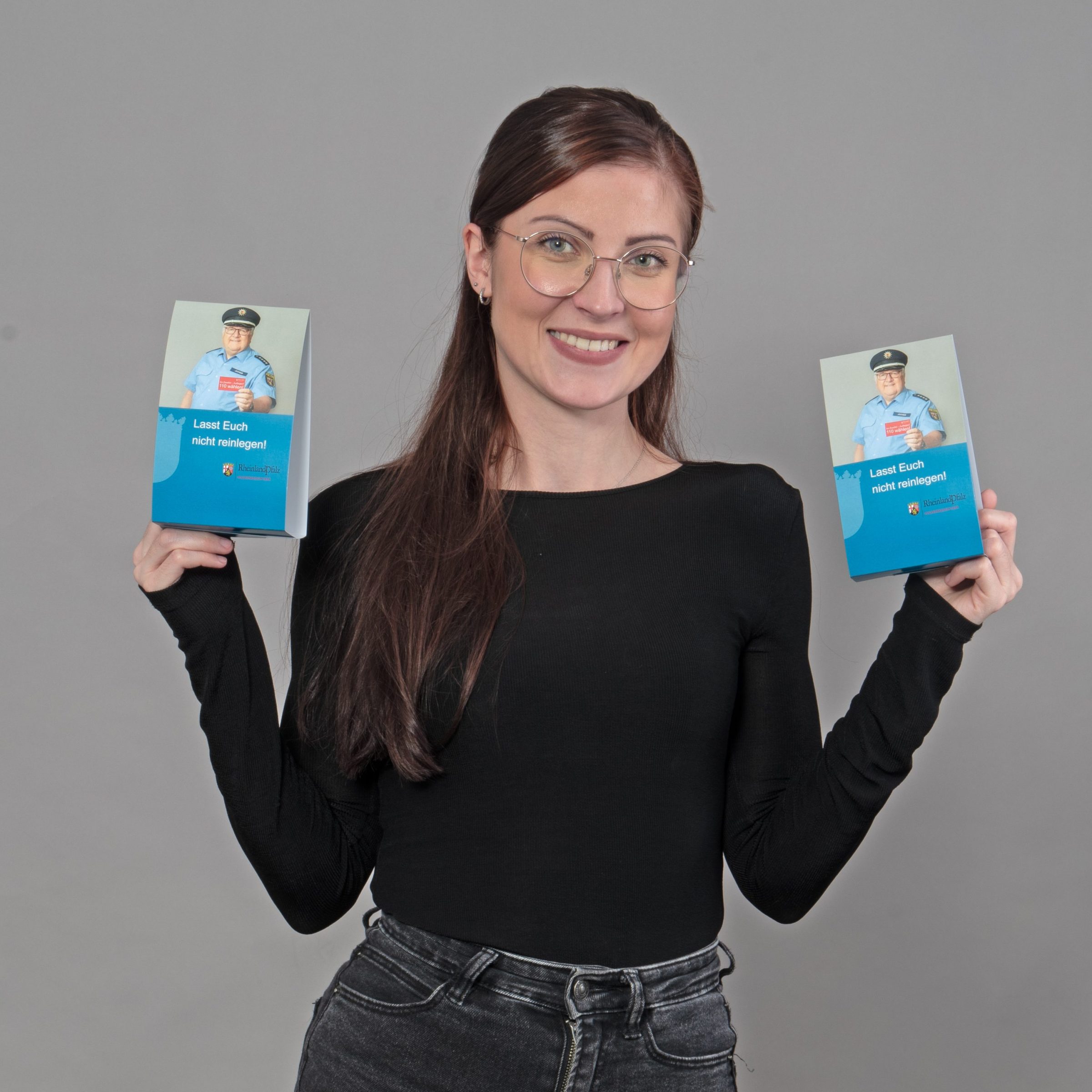Frau hält zwei Aufsteller in den Händen, die zwei Polizisten abbilden, der eine Karte in die Kamera hält, auf der "Im Zweifelsfall 110 wählen" steht. Als Titel des Aufstellers unter dem Foto steht "Lasst Euch nicht reinlegen!"