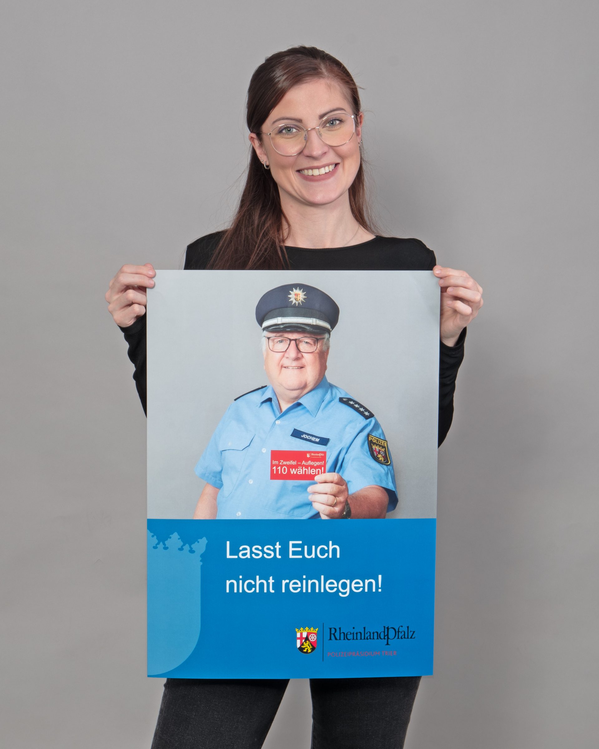 Frau hält ein Plakat, das einen Polizisten abbildet, der eine Karte in die Kamera hält, auf der "Im Zweifelsfall 110 wählen" steht. Als Titel des Plakates unter dem Foto steht "Lasst Euch nicht reinlegen!"