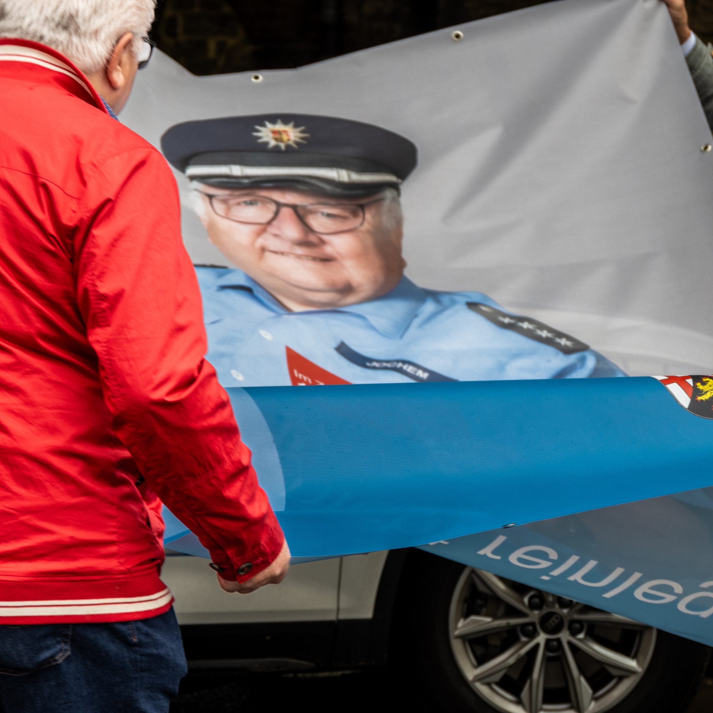 Ein Polizist spannt ein groß bedrucktes Plakat, das einen Polizisten abbildet, der eine Karte in die Kamera hält, auf der "Im Zweifelsfall 110 wählen" steht. Als Titel des Plakates unter dem Foto steht "Lasst Euch nicht reinlegen!"